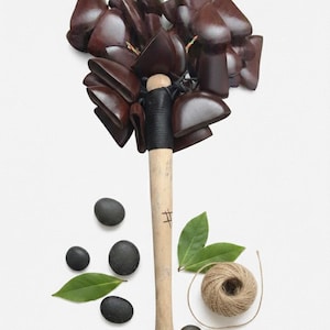 Puede incluir: Un sonajero de campana hecho a mano con un mango de madera y un grupo de campanas marrones. El mango está envuelto en material negro. La imagen también incluye piedras negras, hojas verdes y una bola de hilo. El texto "Handmade Bell Shaker" está en la parte superior.