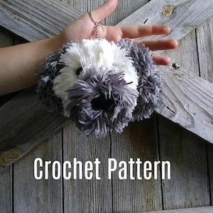 May include: A crochet pattern for a fluffy gray and white dog keychain. The keychain is attached to a silver chain and is held in a hand.
