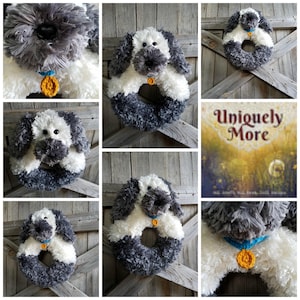 May include: A gray and white crocheted dog wreath with a blue collar and a gold charm. The wreath is hanging on a wooden door with a sign that says "Uniquely More".