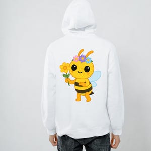 May include: White hoodie featuring a cartoon bee graphic on the back. The bee is yellow and black, holding a flower, and wearing a flower crown. The hoodie has a hood and a drawstring.