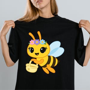 May include: Black t-shirt featuring a cartoon bee with a flower crown, holding a basket. The bee is yellow with black stripes and blue wings. The design is centered on the shirt.