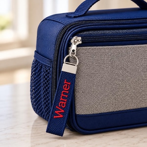 May include: A navy blue lunch bag with a gray front panel, a side mesh pocket, and a top handle. A navy blue fabric zipper pull is attached to the silver metal zipper, featuring the name Warner embroidered in red thread on the fabric strap.
