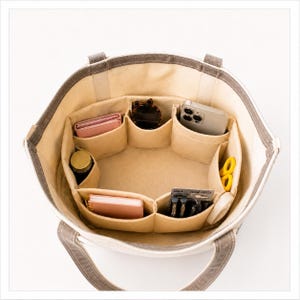 May include: A beige canvas tote bag featuring a structured interior organizer with multiple compartments. The organizer holds a pink wallet, sunglasses, a smartphone, a small notebook, pens, yellow scissors, and a small cylindrical container.