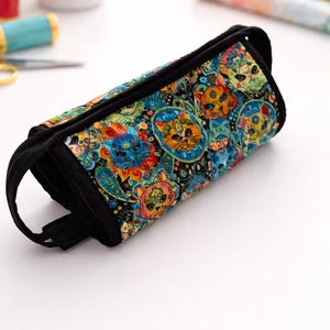 May include: A colorful pencil case featuring a cat-themed pattern in shades of blue, orange, and yellow on a black background. The case has a black zipper and a black handle. The case is approximately 8 inches long.