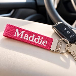 May include: A hot pink fabric keychain strap featuring the white embroidered name Maddie. The strap is attached to a silver-toned metal clasp and a split ring, which holds a black car key fob and a standard silver-colored metal house key.