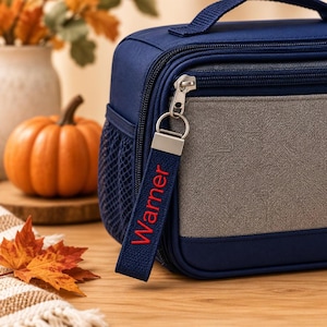 May include: A navy blue lunch bag with a gray front panel, a side mesh pocket, and a top handle. A navy blue fabric zipper pull is attached to the silver zipper, featuring the name Warner embroidered in red thread on the strap.