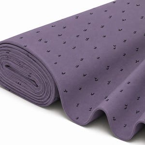 May include: A roll of purple fabric featuring a repeating pattern of small, black, V-shaped marks. The textile is wound into a tight cylinder on one side, with the remaining length of the material draped in soft, flowing folds across the surface.