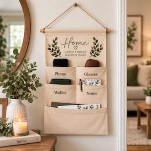 May include: A beige canvas wall organizer with a wooden dowel and hanging cord. The fabric features green leaf illustrations and the text "Home GOOD THINGS HAPPEN HERE". It has six pockets labeled "Phone", "Glasses", "Wallet", and "Notes".