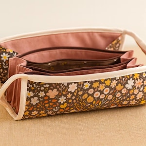 May include: A floral print purse with a handle. The purse features a brown background with yellow, orange, and white flowers. The interior is a solid pink color. The purse has a cream-colored trim and a handle.