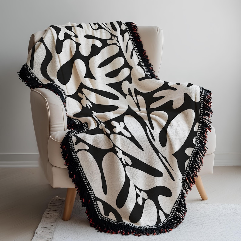 May include: A cream-coloured throw blanket with a bold black abstract pattern is draped over a beige armchair. The blanket has a black fringe with a red accent. The design features organic shapes.