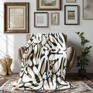 May include: A cream throw blanket with a striking black abstract design, draped over a neutral armchair. The blanket's pattern includes organic shapes and floral motifs. The room features framed artwork and a potted plant.