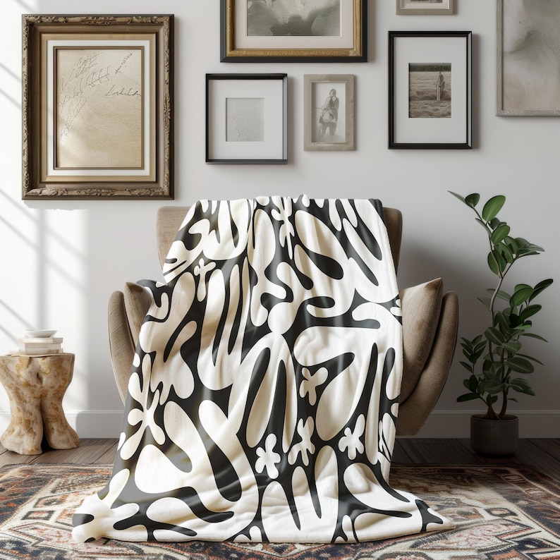 May include: A cream throw blanket with a striking black abstract design, draped over a neutral armchair. The blanket's pattern includes organic shapes and floral motifs. The room features framed artwork and a potted plant.