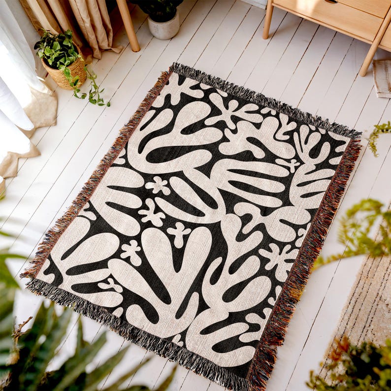 May include: A rectangular woven throw blanket with a black and white abstract pattern. The design features organic shapes and floral elements. The blanket has a fringed edge.