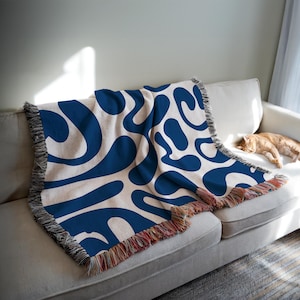May include: A woven throw blanket featuring a cream-coloured background and a repeating pattern of large, abstract blue shapes. The blanket has a fringed edge with a mix of colours. The blanket is draped over a white sofa.