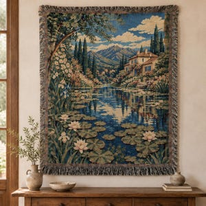 May include: A rectangular woven tapestry wall hanging featuring a landscape scene with a stone house, a calm pond filled with lily pads and blooming water lilies, tall cypress trees, and distant mountains under a blue sky with white clouds.