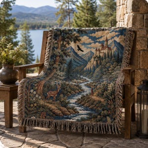 May include: A woven throw blanket featuring a detailed landscape scene with a castle on a mountain, a flowing river, a deer in the foreground, and a bird in flight. The blanket has a fringed edge and displays a palette of blue, green, and brown.