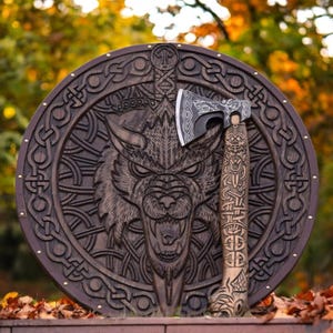 May include: A wooden shield and axe set with intricate Viking-style carvings. The shield features a detailed wolf head design, while the axe has a wolf head on the blade and a carved handle. The set is brown with metallic accents.
