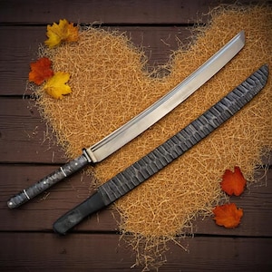 May include: A katana sword with a silver blade and a textured black scabbard. The handle is dark with silver accents. The sword is displayed on a bed of straw with autumn leaves.