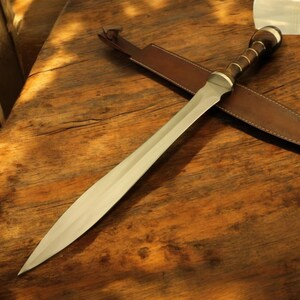 Könnte beinhalten: Ein silbernes Schwert mit einem braunen Ledergriff und einer braunen Lederscheide. Das Schwert liegt auf einer Holzoberfläche.