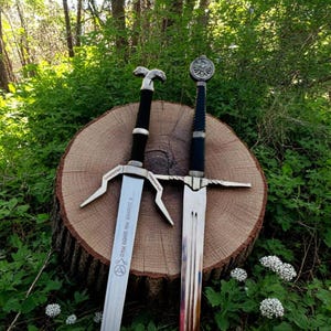 May include: Two silver swords with black handles and intricate details. The swords are resting on a wooden stump in a green grassy area. The sword on the left has the inscription "A LUNDWIGE FAN DORDJA PRAND" on the blade.