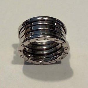 May include: A silver-toned metal ring with a multi-band, tiered design. The outer edges of the ring feature the engraved text BVLGARI. The surface is polished and reflective, showing a cylindrical shape with a hollow center.