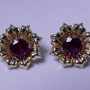 May include: A pair of gold flower-shaped earrings with a large, round, dark purple gemstone in the center of each flower.