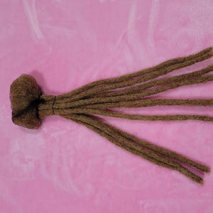 May include: A bundle of brown dreadlocks tied together with a black band. The dreadlocks are arranged in a fan shape, with several individual locks extending outwards. The background is a solid pink color.