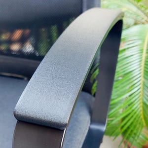 Puede incluir: Primer plano de un reposabrazos negro de silla de oficina. El reposabrazos tiene una parte superior curva y acolchada en color gris oscuro, con una superficie texturizada. El marco es negro y tiene un acabado liso y brillante. El respaldo de malla de la silla es visible.