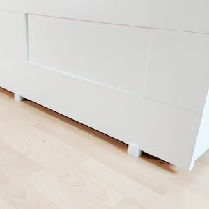 May include: A white cabinet with a clean, modern design. The cabinet has a rectangular shape and is supported by short, cylindrical legs. The surface is smooth and the color is a bright white. The cabinet is positioned on a light-colored wooden floor.
