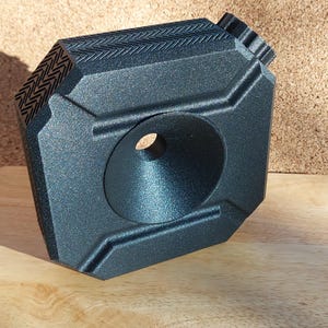 May include: A black, geometric-shaped object with a textured surface. It features a large, circular opening and a decorative chevron pattern on one side. The object is made of a matte material and sits on a wooden surface.