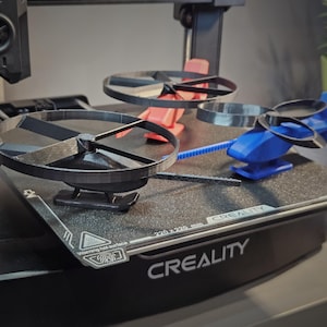 May include: A 3D printer displaying several miniature helicopter models in various stages of completion. The models are black and blue, with red support structures. The printer bed has the text "CREALITY" and "220x220 mm".