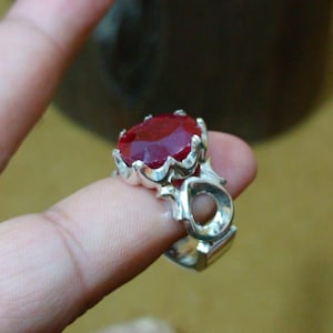 May include: A silver ring with a large, oval-cut, deep red gemstone. The ring has an ornate, detailed setting with a floral design. The gemstone is the focal point, catching the light and adding a touch of elegance.