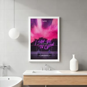Op de afbeelding: Een ingelijste poster met de tekst "I SAW THE TV GLOW" in paars neon tegen een roze en zwarte achtergrond. De poster toont twee figuren en is geplaatst in een witte lijst.