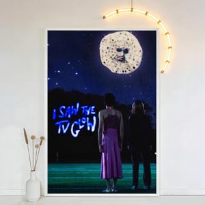 Op de afbeelding: Een ingelijste kunstdruk met een surrealistische nachtsc&egrave;ne. Twee figuren staan op een veld en kijken naar een maanachtig object in de lucht. De tekst "I SAW THE TV GLOW" is blauw verlicht. Het kunstwerk heeft een vintage, dromerige kwaliteit.