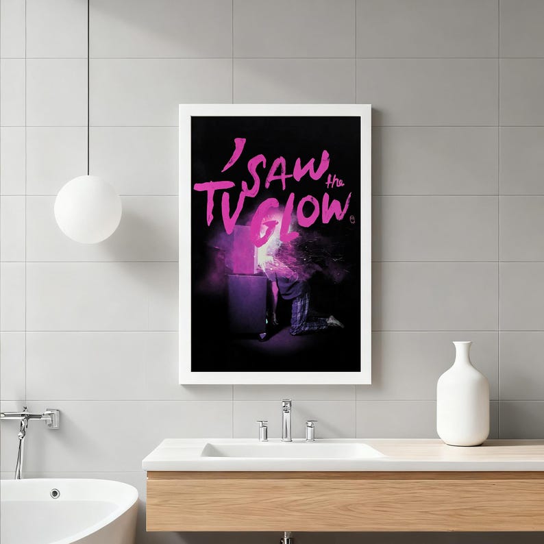 Op de afbeelding: Een ingelijste kunstdruk met de tekst "I Saw the TV Glow" in roze. Het kunstwerk toont een persoon die knielt voor een gloeiende televisie. De achtergrond is zwart en de afbeelding heeft een surrealistische, dromerige kwaliteit.