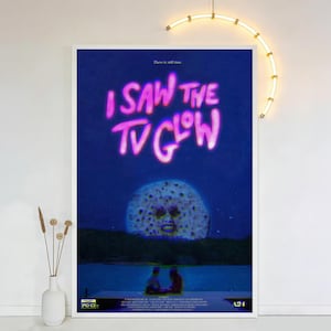 Op de afbeelding: Een ingelijste poster met de tekst "I SAW THE TV GLOW" in roze, tegen een donkerblauwe achtergrond. De poster toont een surrealistisch beeld van een maan met een gezicht en twee figuren op een steiger. De poster wordt op een witte muur getoond.