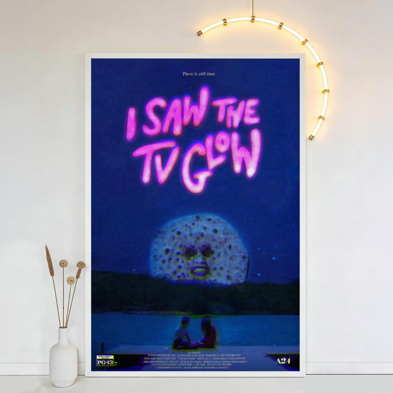 Op de afbeelding: Een ingelijste poster met de tekst "I SAW THE TV GLOW" in roze, tegen een donkerblauwe achtergrond. De poster toont een surrealistisch beeld van een maan met een gezicht en twee figuren op een steiger. De poster wordt op een witte muur getoond.