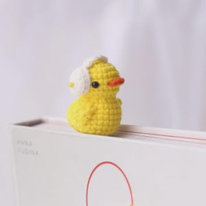 May include: A small, yellow crocheted duck figurine wearing white earmuffs. The duck has a bright orange beak and black eyes. The figurine is sitting on top of a white book with the name "ANNA YUDINA" printed on the side.