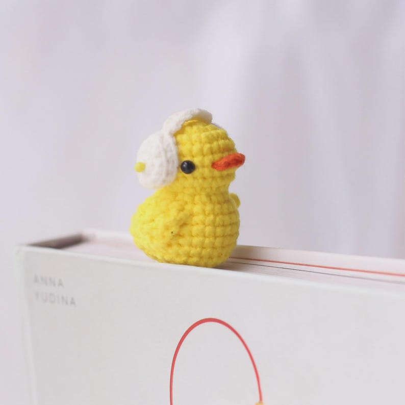 May include: A small, yellow crocheted duck figurine wearing white earmuffs. The duck has a bright orange beak and black eyes. The figurine is sitting on top of a white book with the name "ANNA YUDINA" printed on the side.