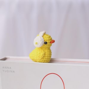 May include: A small, handmade yellow crochet duck wearing white earmuffs. The duck has a small orange beak and black eyes. The duck is sitting on a white book with the name "ANNA YUDINA" printed on the side.