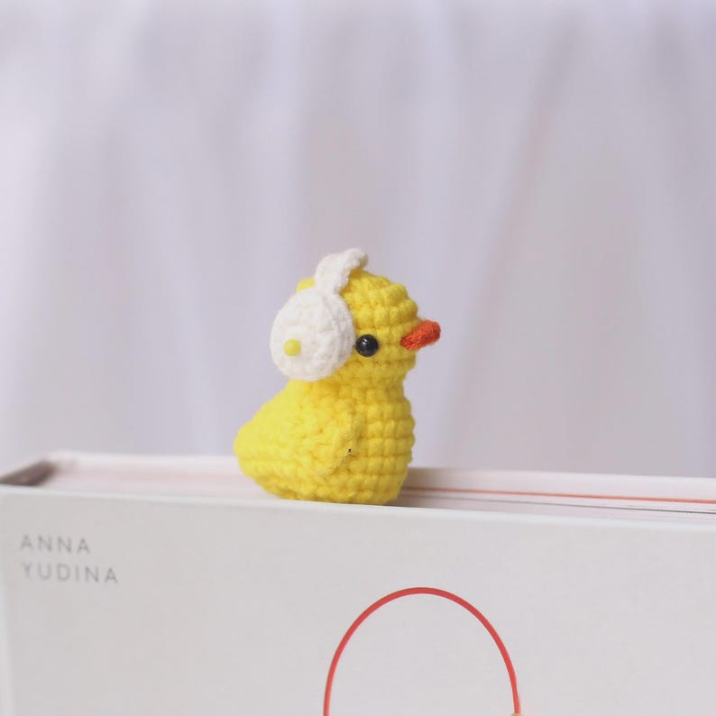 May include: A small, handmade yellow crochet duck wearing white earmuffs. The duck has a small orange beak and black eyes. The duck is sitting on a white book with the name "ANNA YUDINA" printed on the side.