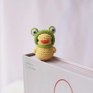 May include: A handmade crochet duckling wearing a green frog hat, perched on a book. The duckling is yellow with an orange beak and small black eyes. The frog hat has large, white-eyed details. The book has the name "ANNA YUDINA" printed on it.