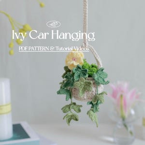 以下が含まれることがあります： 黄色の花と緑のアイビーの葉が付いた、かぎ針編みで作った吊り下げ式のプランター。プランターは白い糸でできており、茶色のロープで吊り下げられています。画像には「Ivy Car Hanging」と「PDF Pattern & Tutorial Videos」のテキストが表示されています。