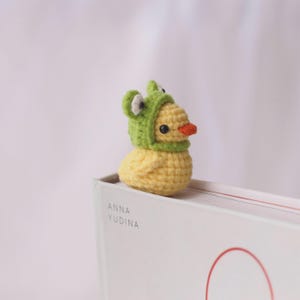May include: A small, yellow crocheted duck wearing a green frog hat with white eyes and an orange beak. The duck is sitting on a white book with the text "ANNA YUDINA" printed on the side. The background is a soft, blurred white.