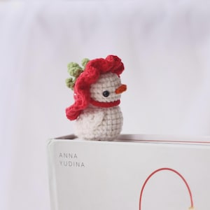 May include: A small, crocheted snowman figurine. The snowman is white with a red scarf and hat, a small orange carrot nose, and black eyes. The snowman is approximately 2 inches tall. The figurine is sitting on a white surface.