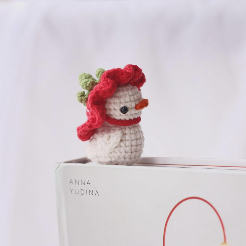 May include: A small, crocheted snowman figurine. The snowman is white with a red scarf and hat, a small orange carrot nose, and black eyes. The snowman is approximately 2 inches tall. The figurine is sitting on a white surface.