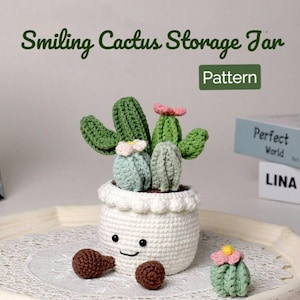 May include: Smiling cactus storage jar crochet pattern. A white crocheted jar with a smiling face holds a group of green crocheted cacti with pink flowers. The jar is sitting on a white tray with a lace doily.