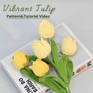 Può includere: Un mazzo di tulipani gialli all'uncinetto con steli e foglie verdi. I tulipani sono disposti su un libro bianco con il testo "BABADI" visibile.