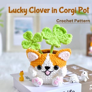 May include: A crocheted lucky clover plant in a crocheted corgi pot. The corgi pot is white with orange and brown accents and has black eyes and a pink nose. The clover plant is green and has four leaves on each stem. The text "Lucky Clover in Corgi Pot" and "Crochet Pattern" is visible in the image.