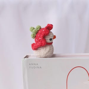 May include: A crocheted snowman figurine with a red hat and green accents. The snowman has a small orange nose and black eyes. The figurine is sitting on a white surface with the text "ANNA YUDINA" visible.
